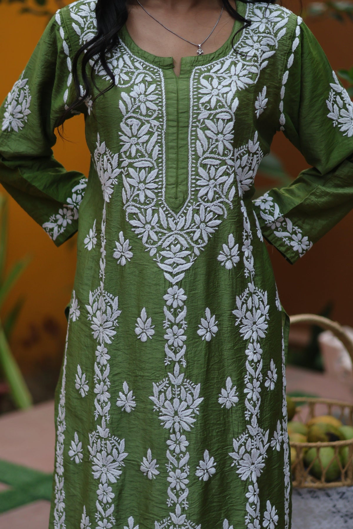 A woman wearing a green long kurta with white floral embroidery and printed patterns, standing against a warm orange-brown background.