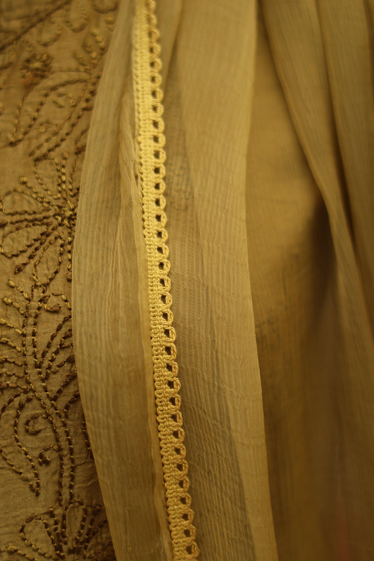 Close-up of a beige fabric garment or drape with a decorative embroidered border and a vertical row of small eyelet cutouts, showing the texture and color of the material.