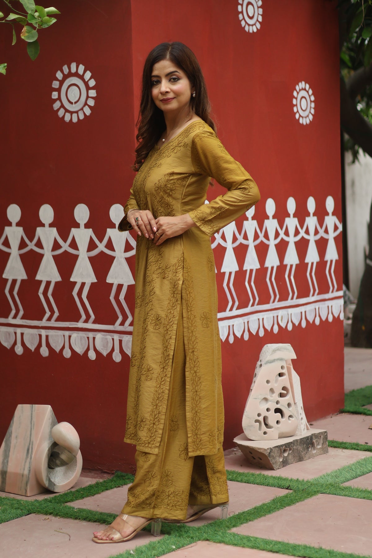 A woman modeling a mustard yellow embroidered kurta and trouser set, standing in front of a red wall decorated with white folk art patterns.