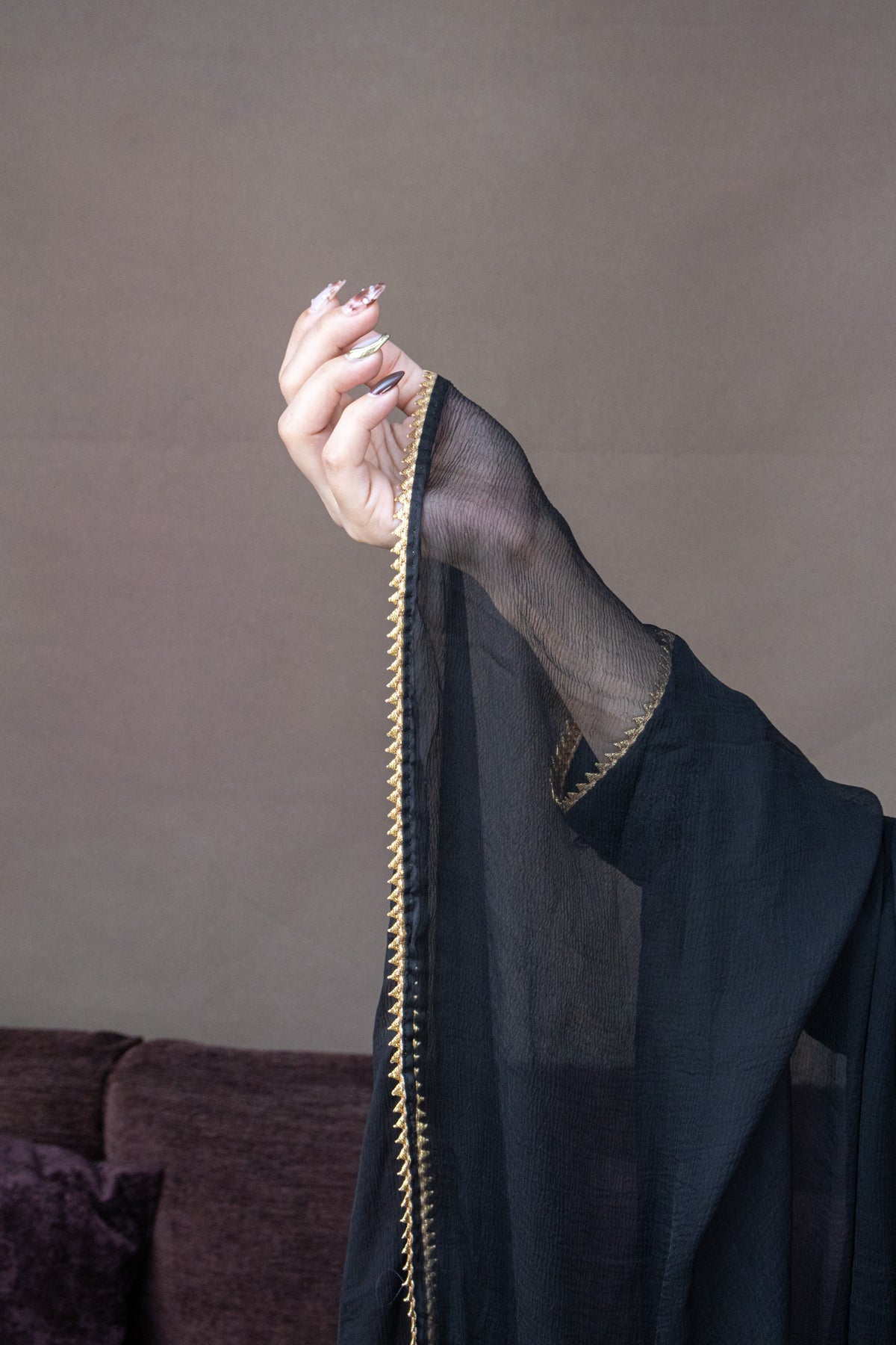 A person's hand and arm wearing a sheer black mesh abaya or long sleeve, held up to show a decorative gold braided trim along the edge, against a plain taupe wall.