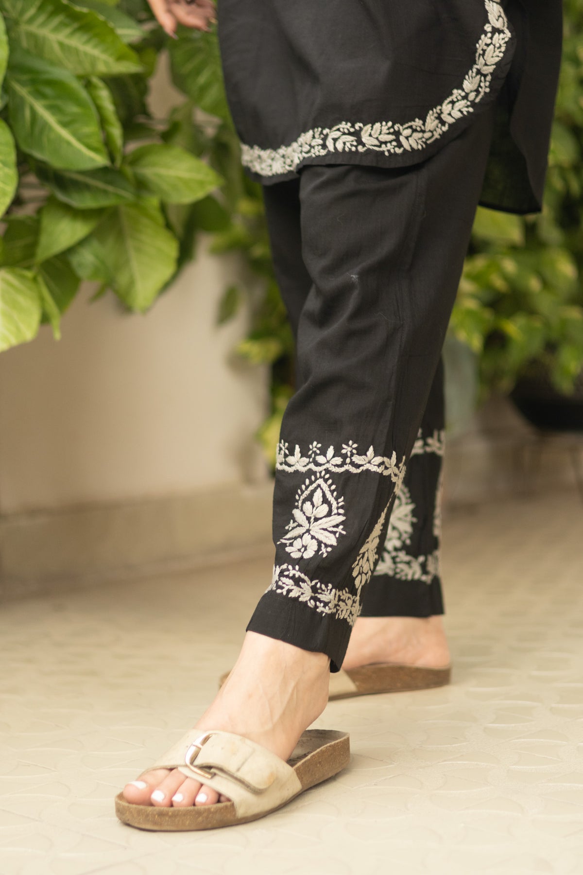 Black pants with white floral embroidery near the hem and ankle, shown on a model wearing beige slide sandals in an indoor setting with green plants in the background.