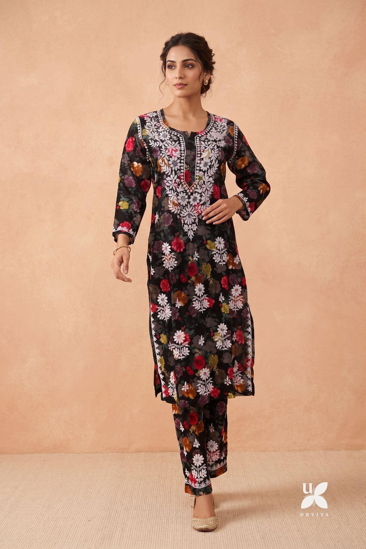 A woman modeling a black three-piece ethnic kurta set with a vibrant multicolor floral print, featuring a long-sleeve tunic, matching trousers, and a detailed embroidered neckline, set against a warm beige studio backdrop. Black Cotton Printed Chikankari Ghaspatti Kurti with Palazzo 2 Piece Set