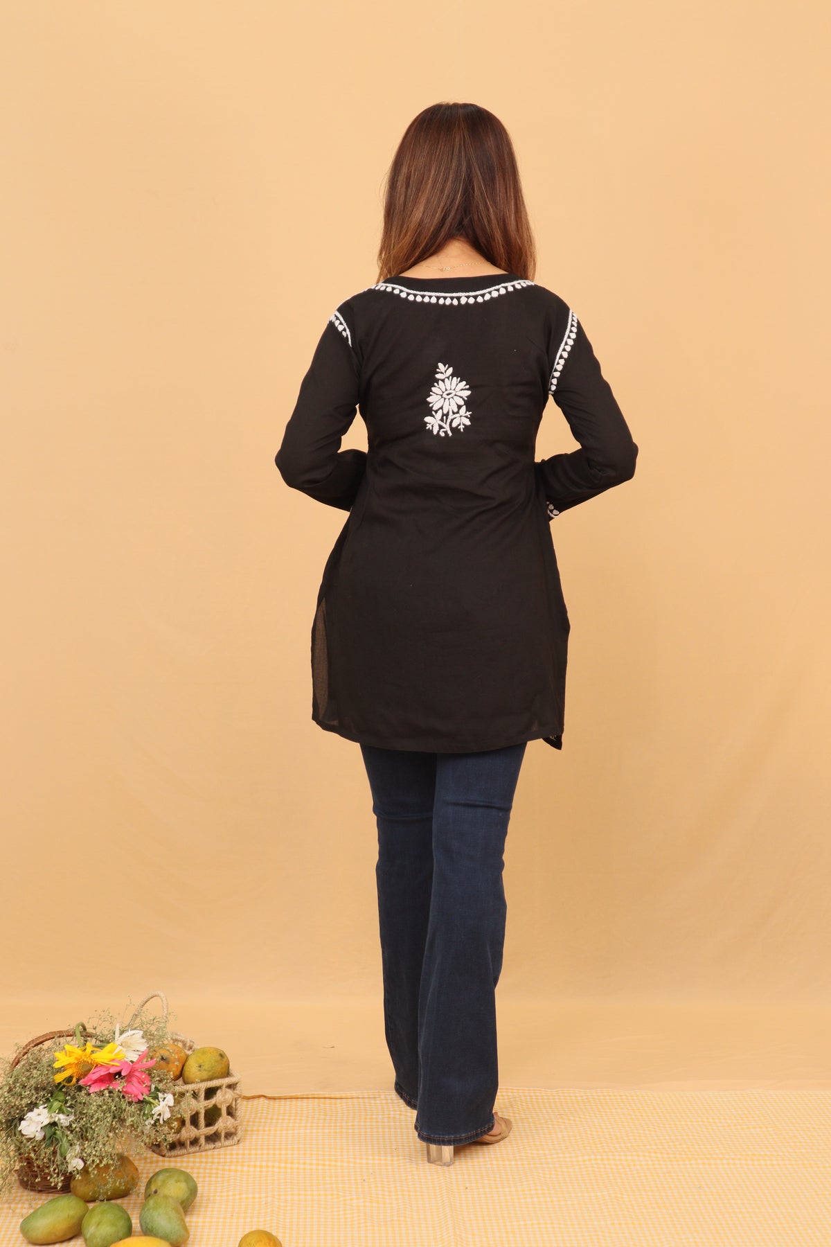 A woman viewed from the back wearing a black long-sleeve kurta with white decorative embroidery on the neckline and upper back, paired with dark blue jeans, standing against a solid beige backdrop.