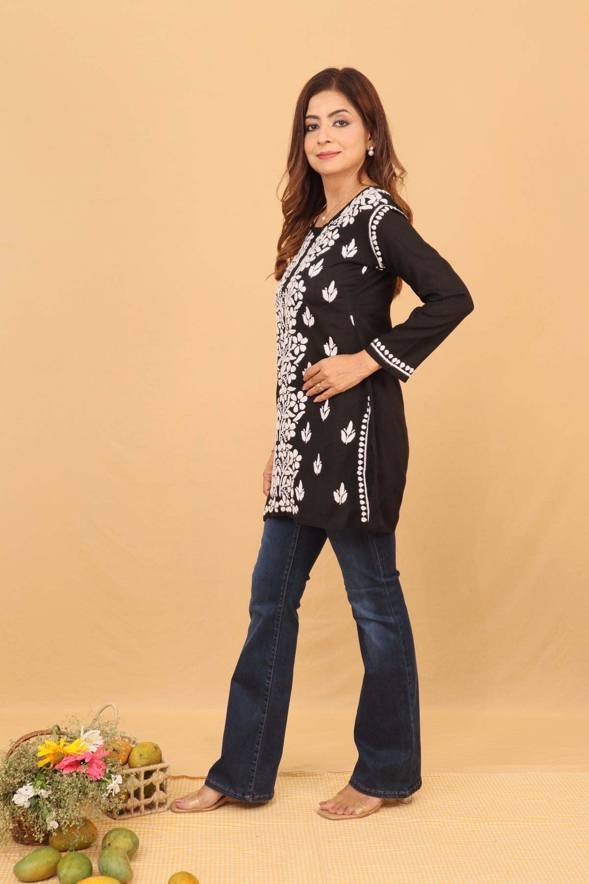 A woman modeling a black long-sleeve kurta with white floral embroidery and silver mirror work, paired with dark blue jeans, against a solid beige backdrop.