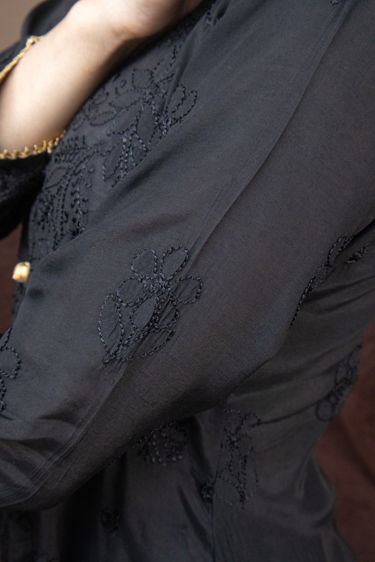 Close-up of a black embroidered garment with floral lace-like detailing and a small gold button, worn by a person whose arm is visible.