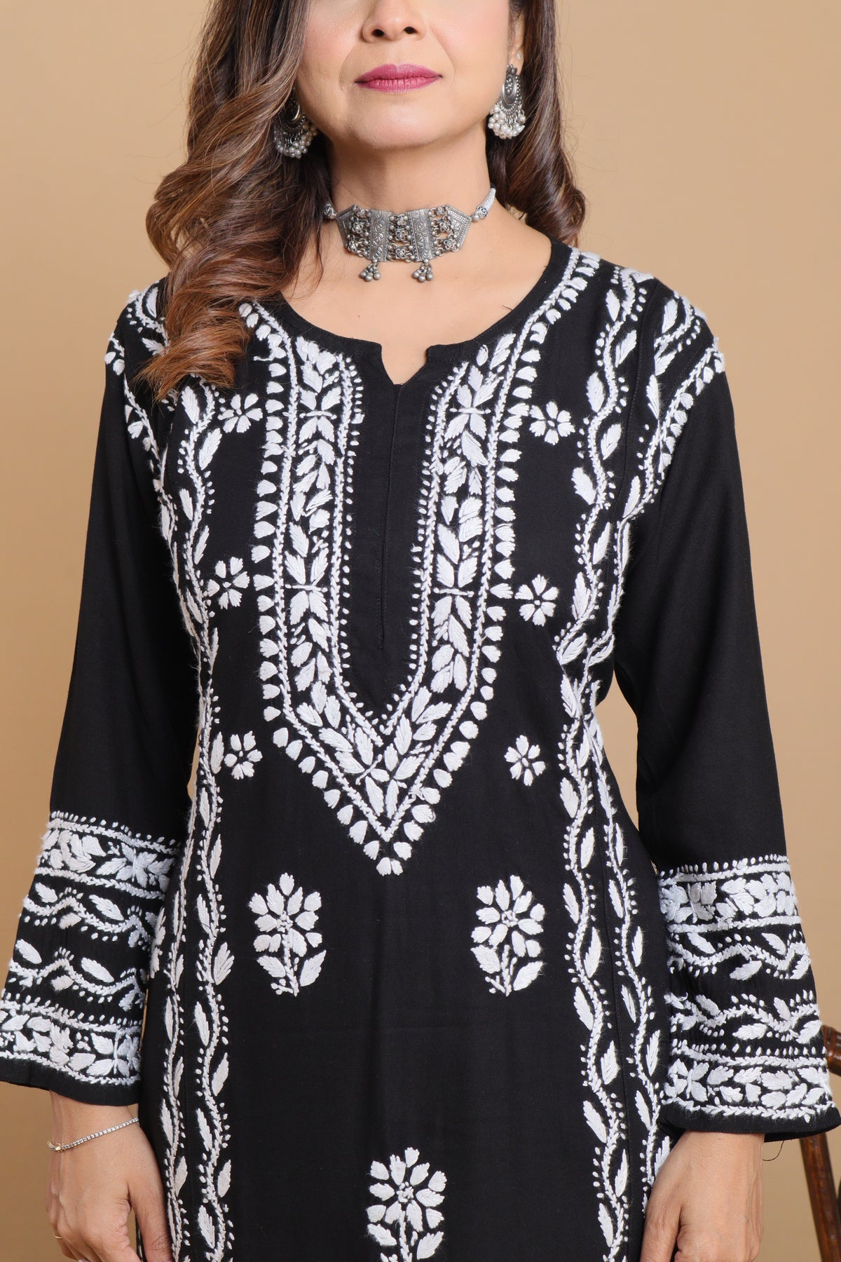 A woman wearing a black long-sleeve kurta with intricate white embroidery, shown against a solid beige background.