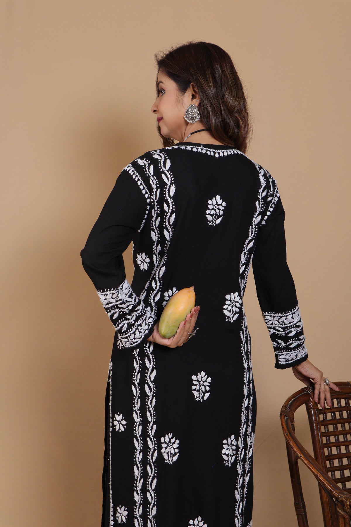 A woman wearing a black long-sleeve kurta with white floral and geometric embroidery, holding a yellow mango against a solid beige background.