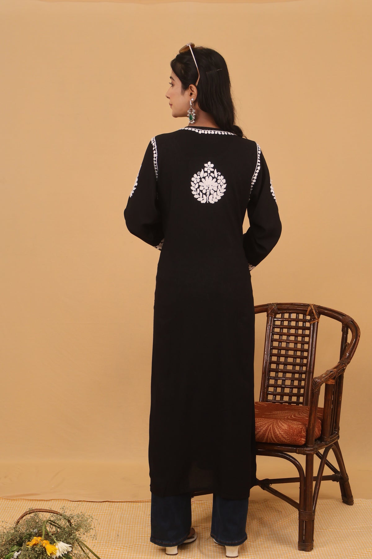 A woman wearing a long black traditional-style dress with white embroidered details and blue pants, standing beside a wooden chair against a plain beige wall.