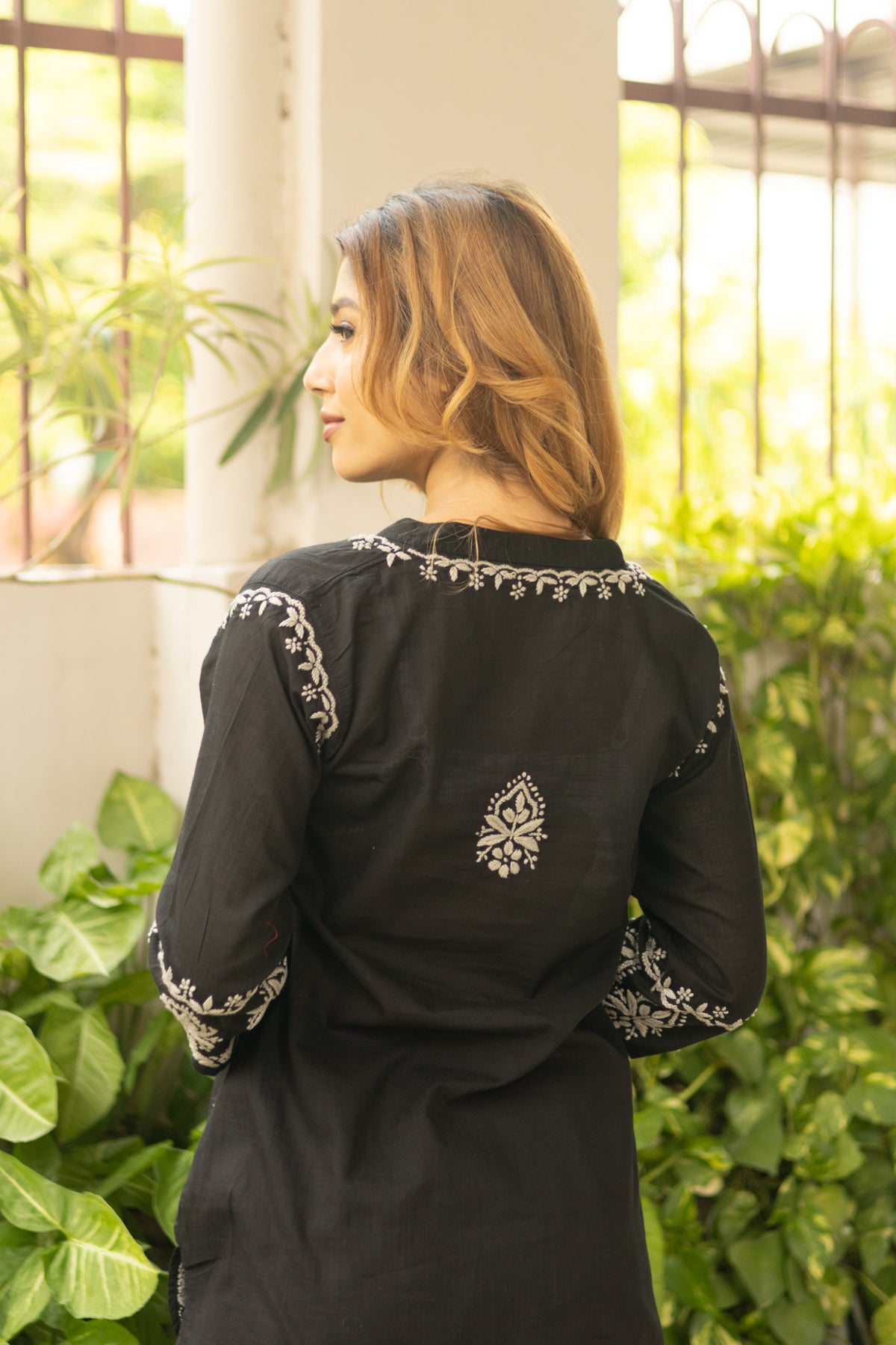 A woman is shown from behind wearing a black long-sleeve kurta with white floral embroidery on the neckline, cuffs, and back. She is standing in a bright, sunlit outdoor patio setting with green plants and white pillars in the background.