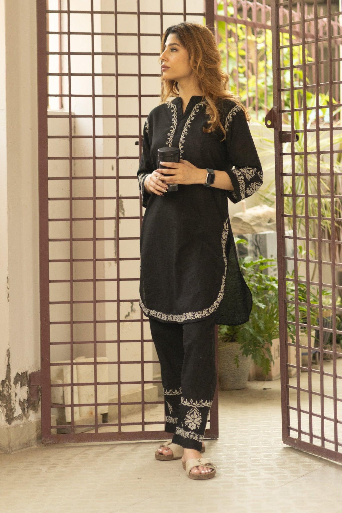 A woman wearing a black three-piece traditional outfit with silver embroidery, standing in a doorway with a metal grid pattern. Black Slub Cotton Chikankari Co-ord Set with White Thread Embroidery