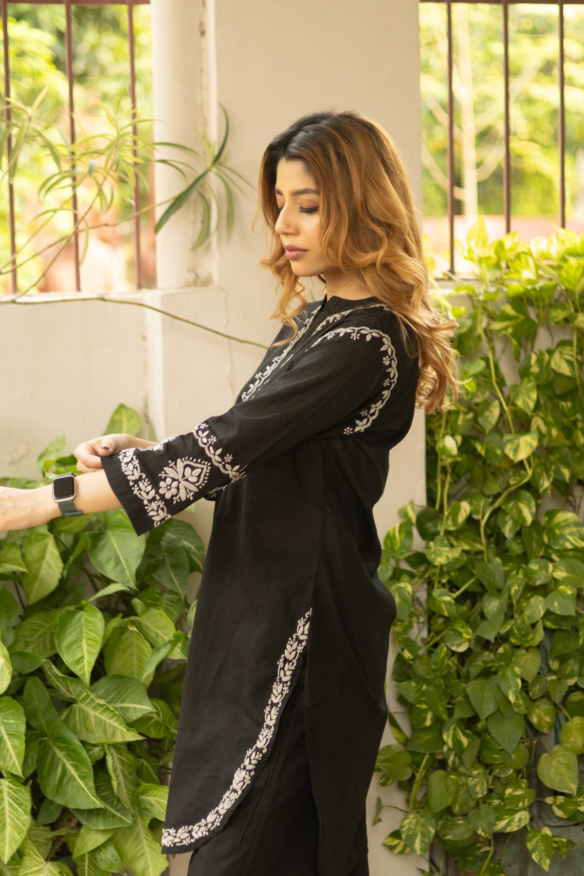 A woman wearing a black long kurta with white floral embroidery on the sleeves and side, standing in front of a wall covered in green plants.
