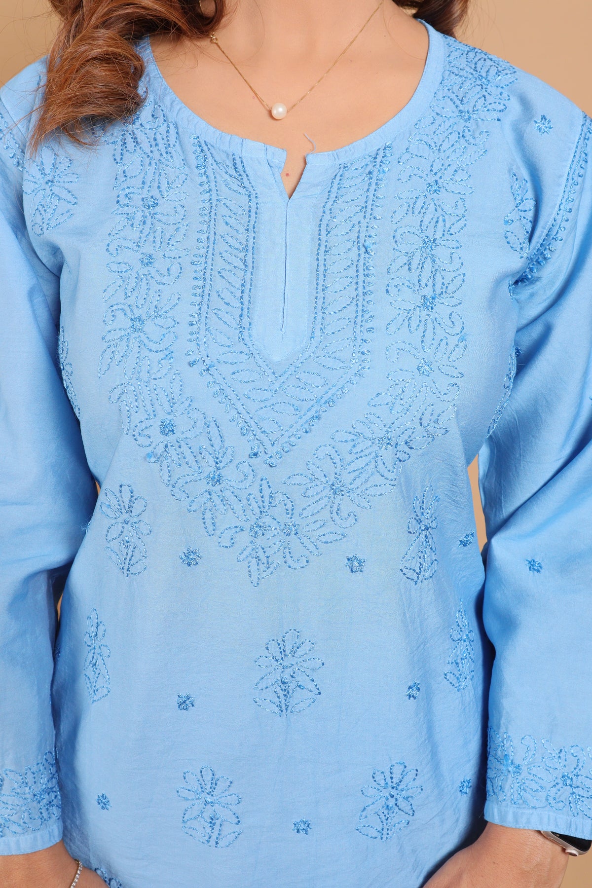 A light blue long-sleeve tunic top with intricate floral embroidery on the chest and cuffs, shown on a model against a solid tan background.