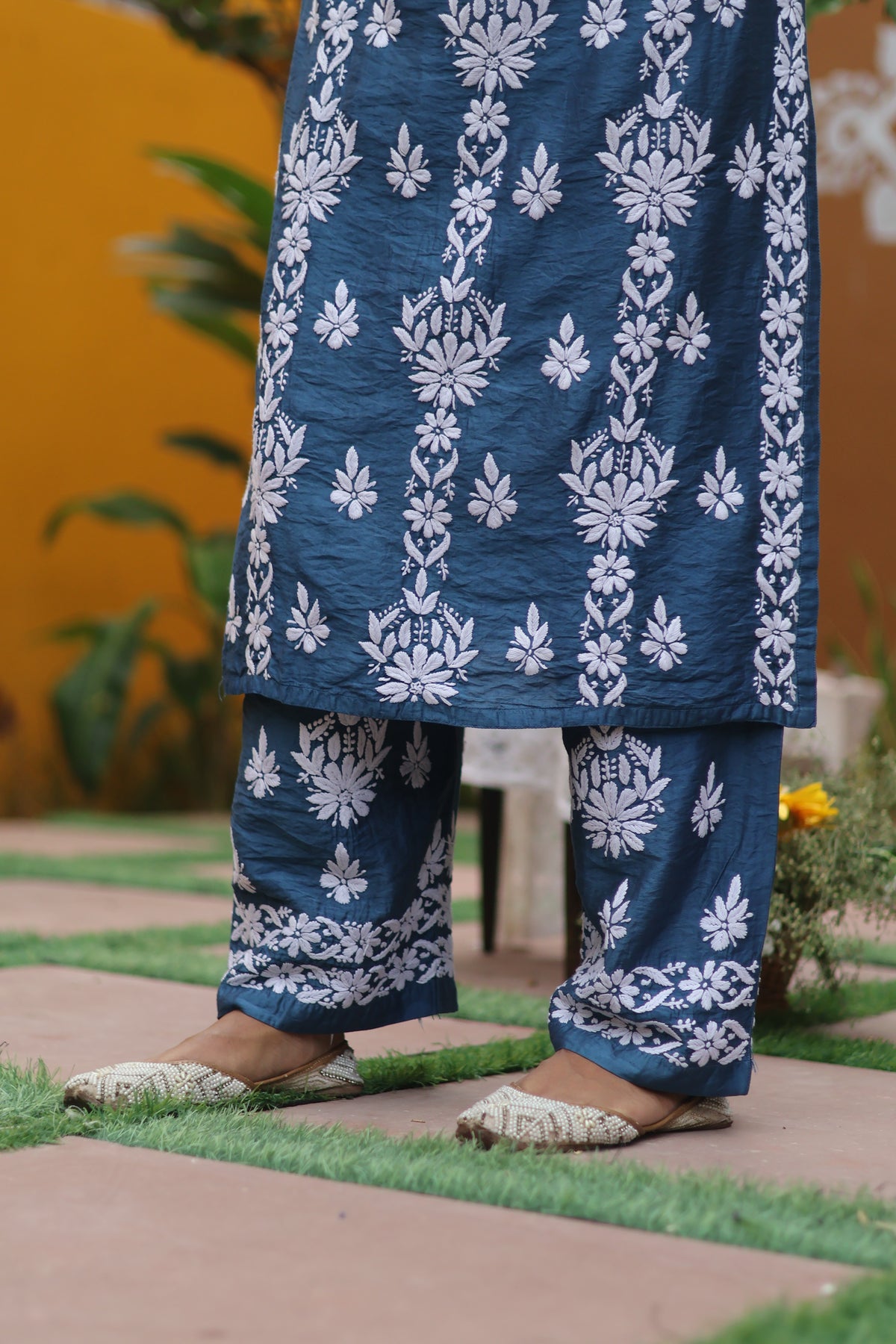A person wearing a navy blue and white printed kurta and matching pants, standing outdoors on a paved path with green grass and a warm yellow-orange wall in the background.