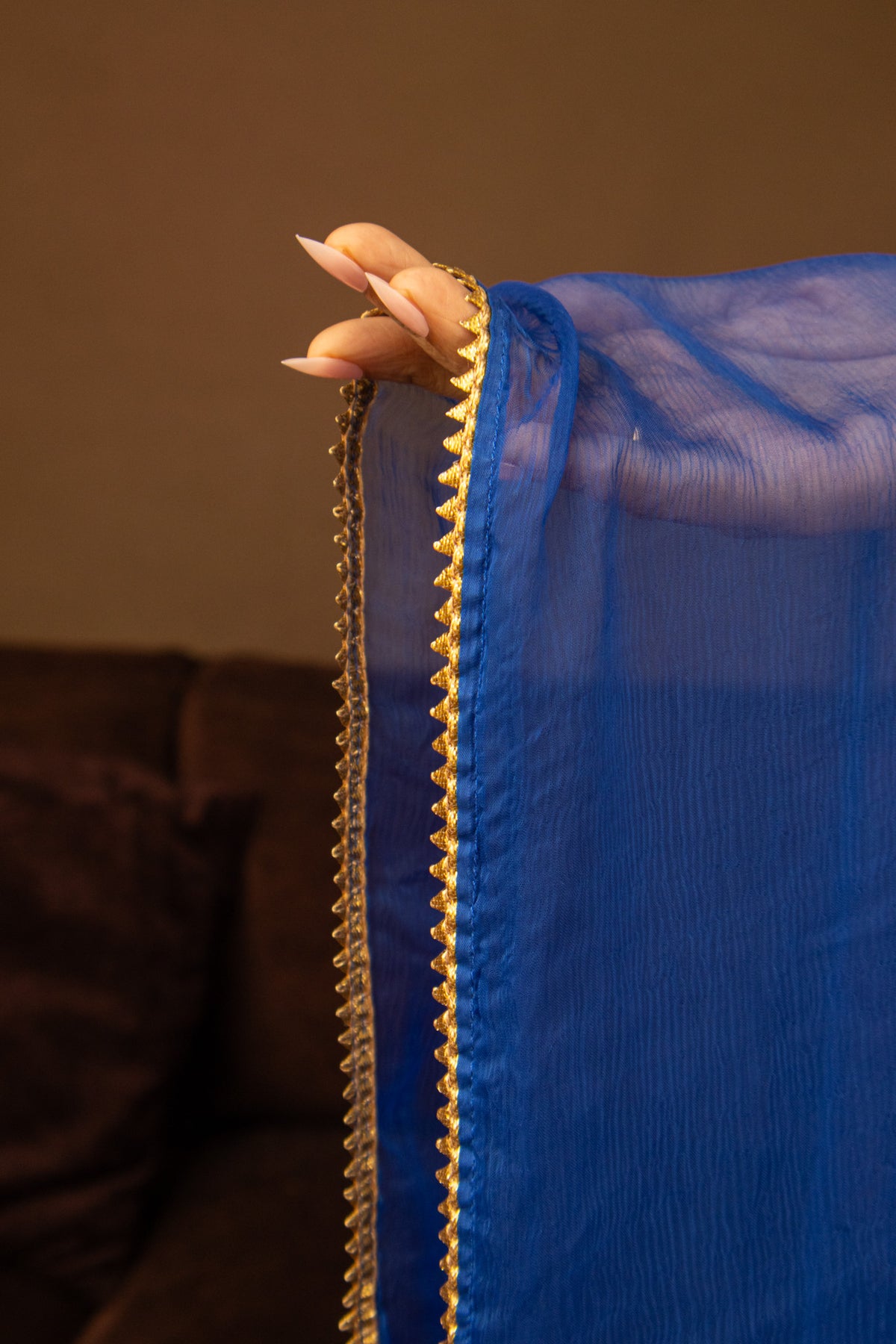 A close-up of a person's hand holding up a vibrant royal blue sheer fabric, likely a dupatta or scarf, featuring a decorative gold metallic zigzag border.