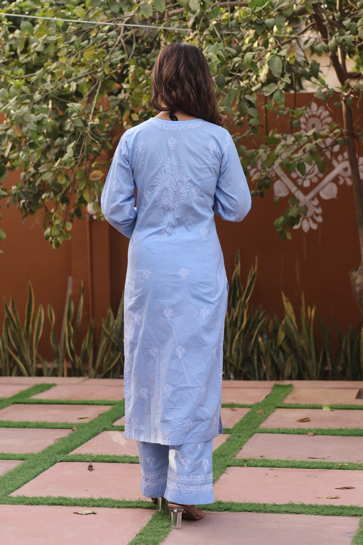 A woman viewed from behind wearing a light blue embroidered kurta and matching trousers in an outdoor garden setting.