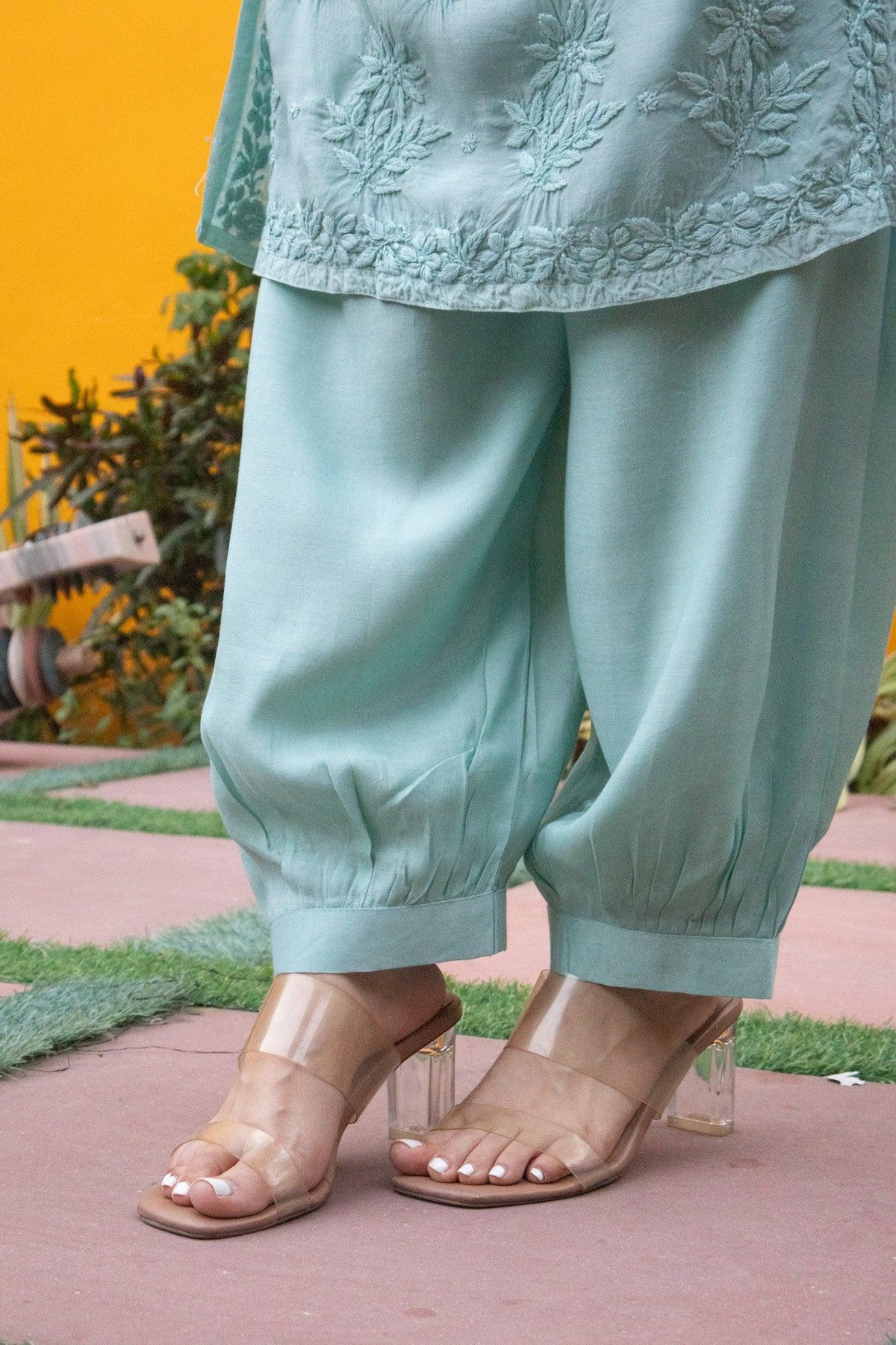 A person wearing a mint green embroidered tunic and matching wide-leg pants with clear high-heel sandals, standing outdoors on a pinkish ground with a yellow wall in the background. Blue Muslin Chikankari Ghaaspatti Long Kurti with Tone-on-Tone Embroidery