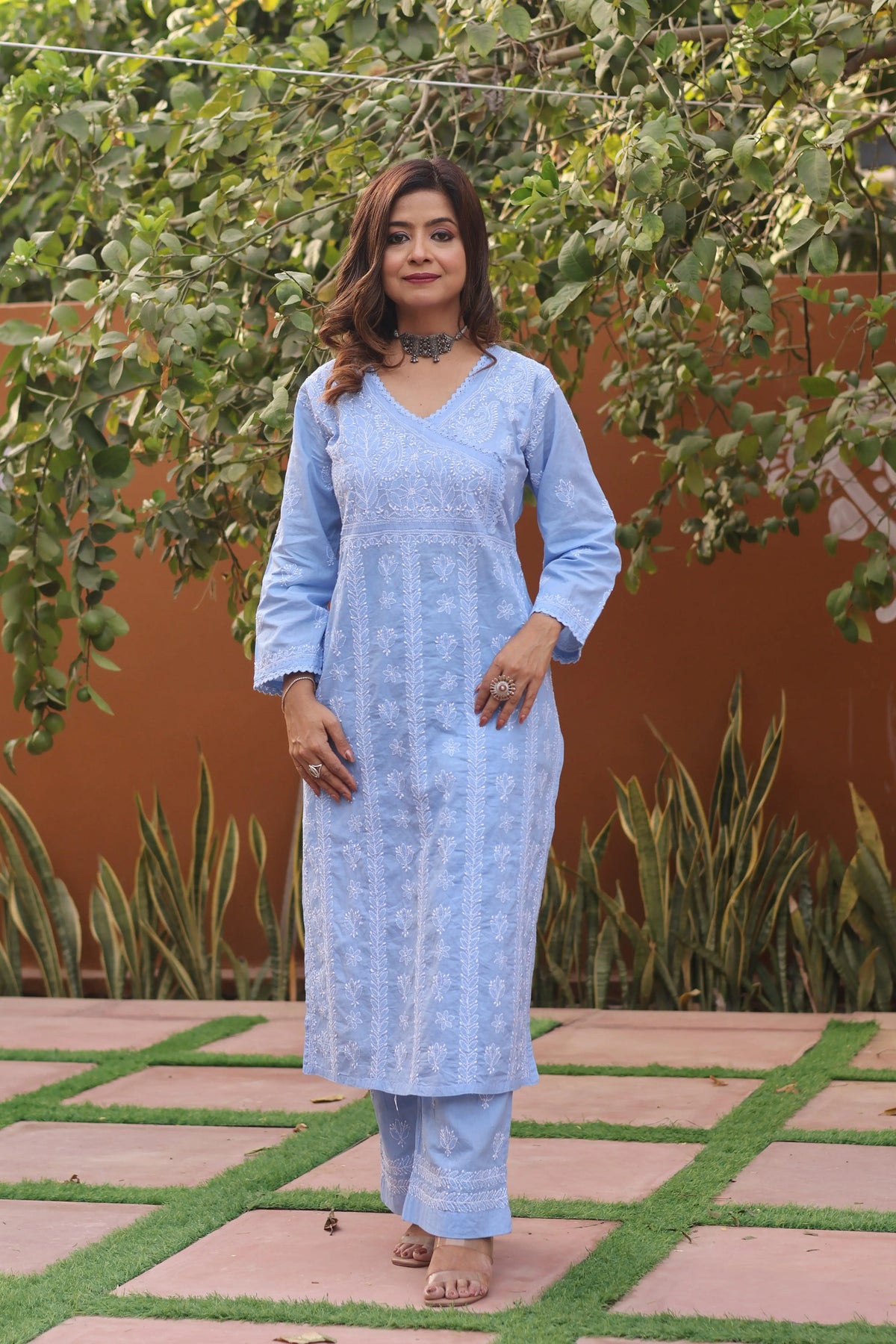 A woman wearing a light blue embroidered kurta and matching pants stands outdoors on a tiled walkway with green plants and a terracotta wall in the background. Blue Cotton Chikankari Angarkha 2 Piece Set Blue Cotton Chikankari Angarkha 2 Piece Set 