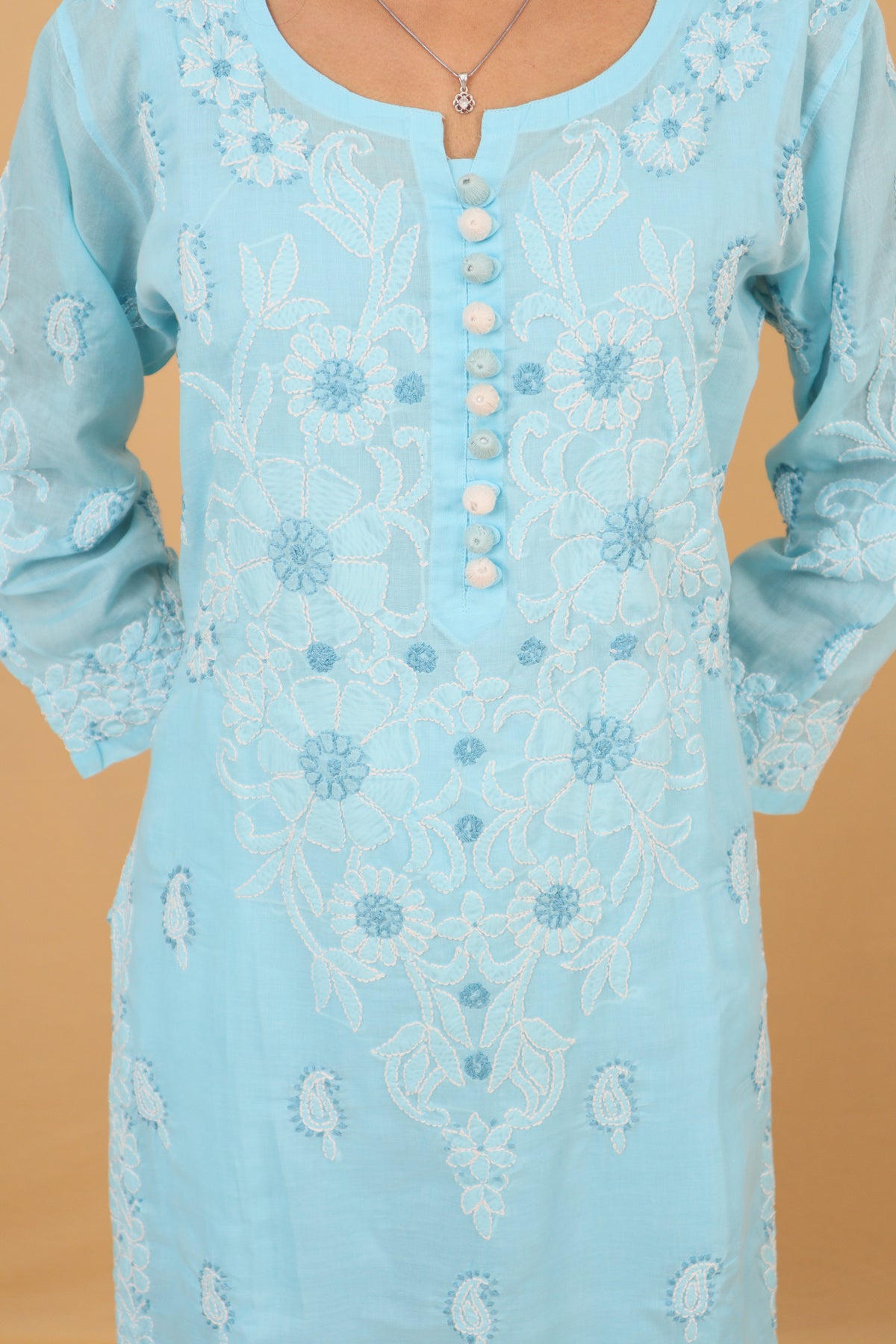 A light blue embroidered tunic with white floral embroidery and a buttoned neckline, shown on a model against a solid tan background.