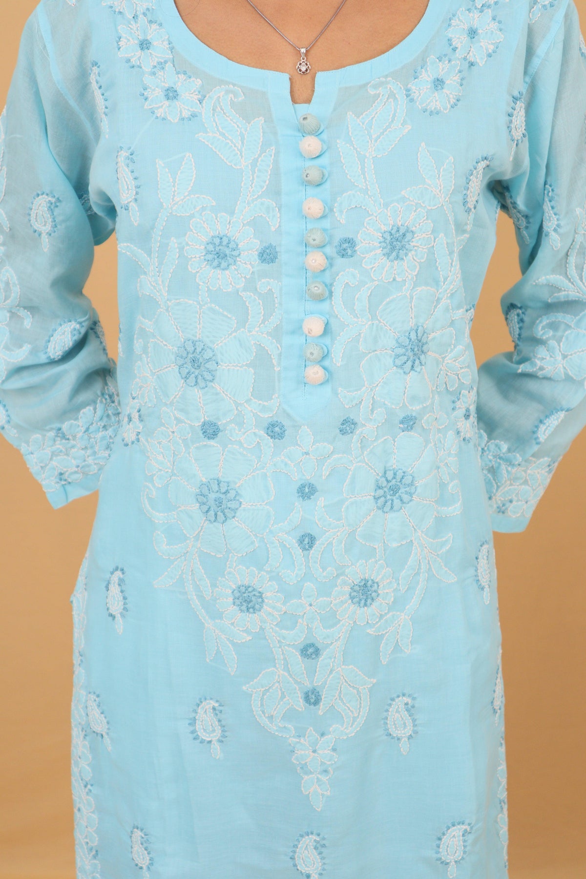 A light blue embroidered tunic with white floral embroidery and a buttoned neckline, shown on a model against a solid tan background. Blue Cotton Chikankari Bakhiya Kurti with White Pants & Dupatta 3 Piece Set
