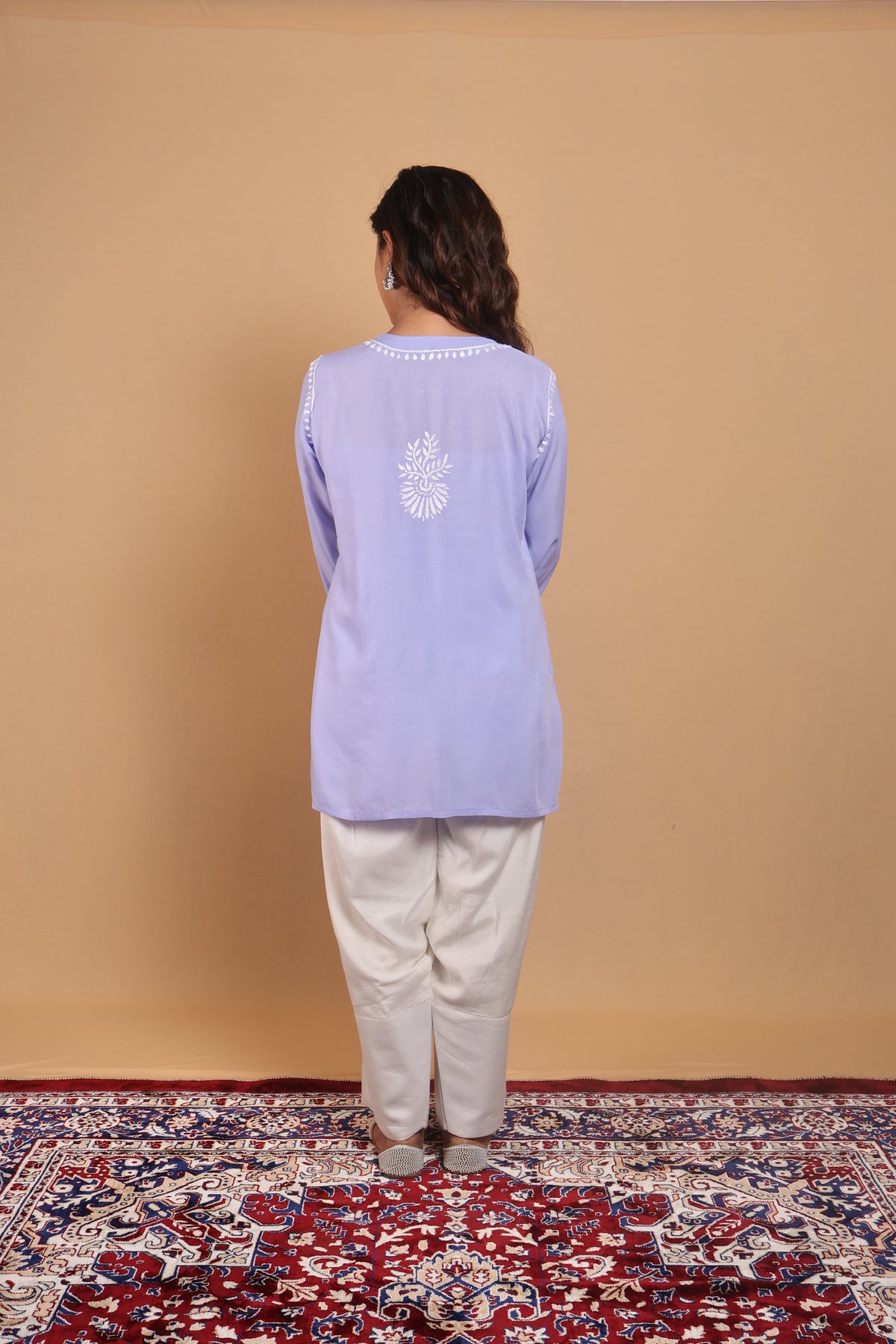A woman stands with her back to the camera wearing a light lavender long-sleeve kurta with white embroidery on the back, paired with off-white trousers, standing on a red and blue patterned rug against a solid beige wall.