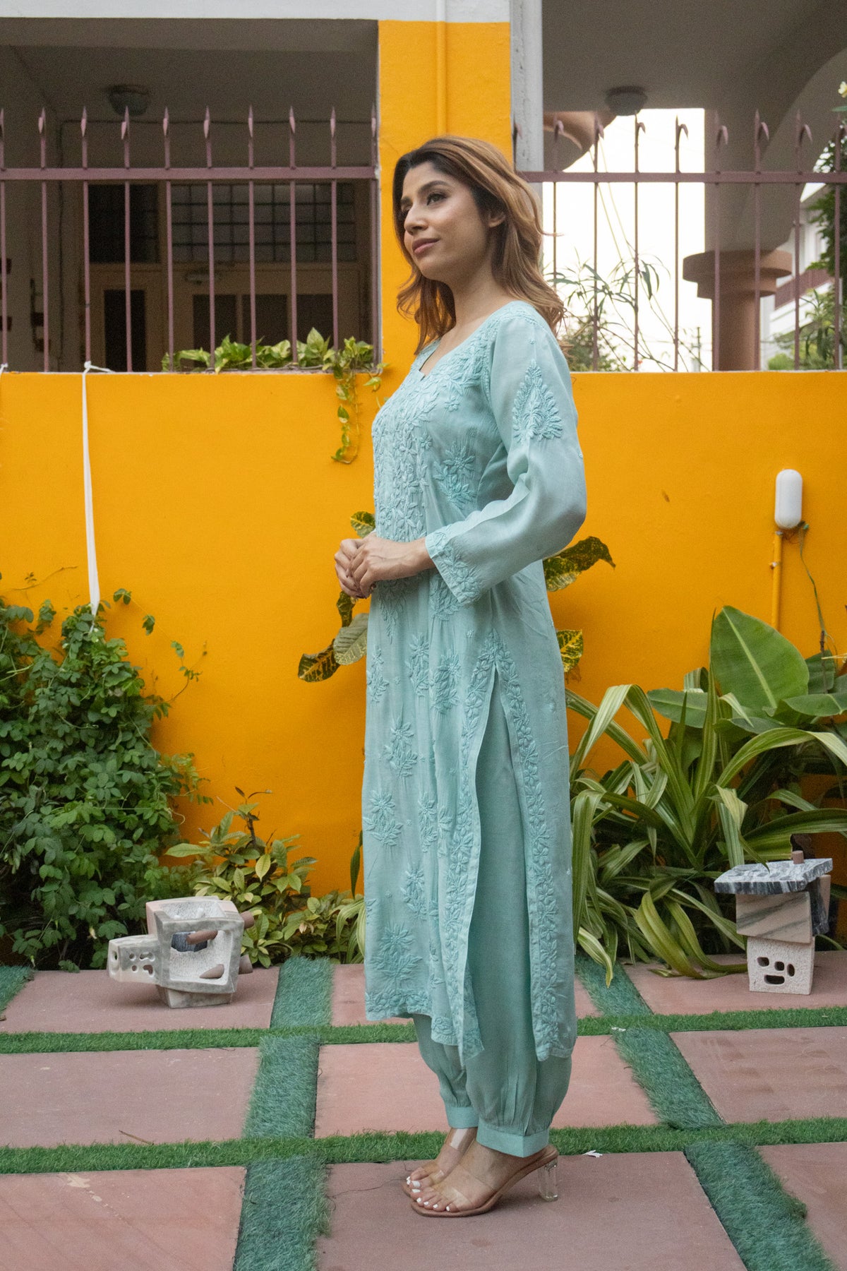 A woman modeling a light blue-green embroidered tunic and trouser set in an outdoor courtyard with a bright yellow wall and green plants.