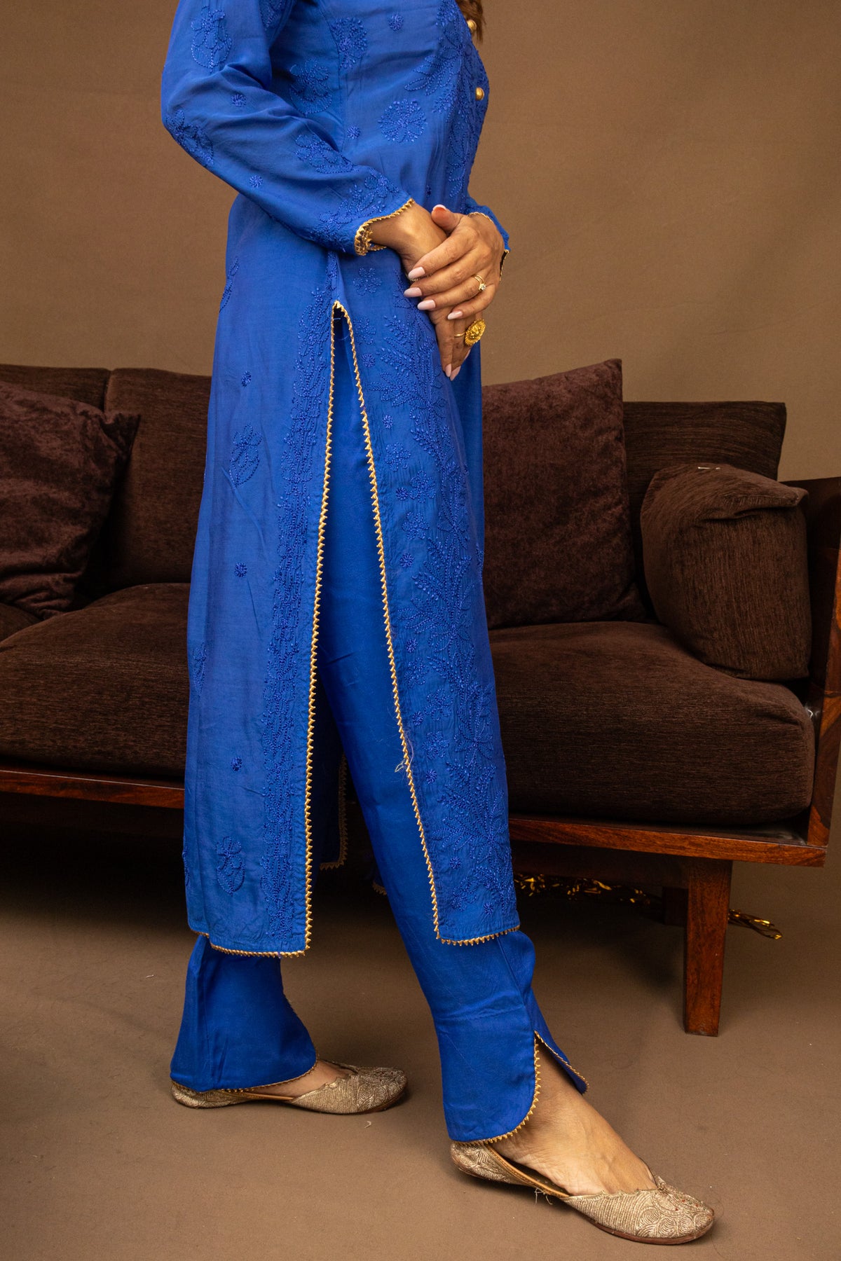 A person wearing a bright royal blue embroidered kurta and matching pants with gold trim, standing indoors in front of a brown sofa. The person is shown from the shoulders down, wearing flat metallic shoes.