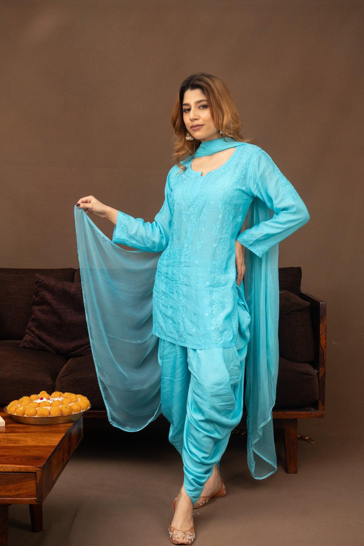 A woman models a light blue traditional South Asian salwar kameez set with a matching dupatta in a studio setting with a brown background and a sofa. Blue Muslin Chikankari Short Kurti with Dhoti and Dupatta 3 Piece Set