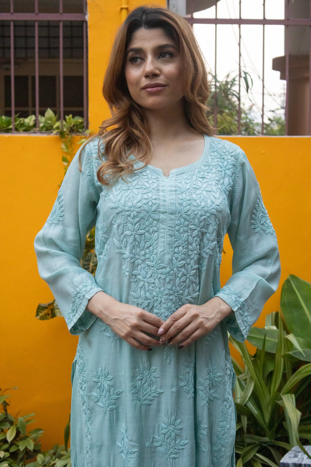A woman wearing a light blue-green embroidered kurta with three-quarter sleeves, standing in front of a bright yellow wall with green plants.
