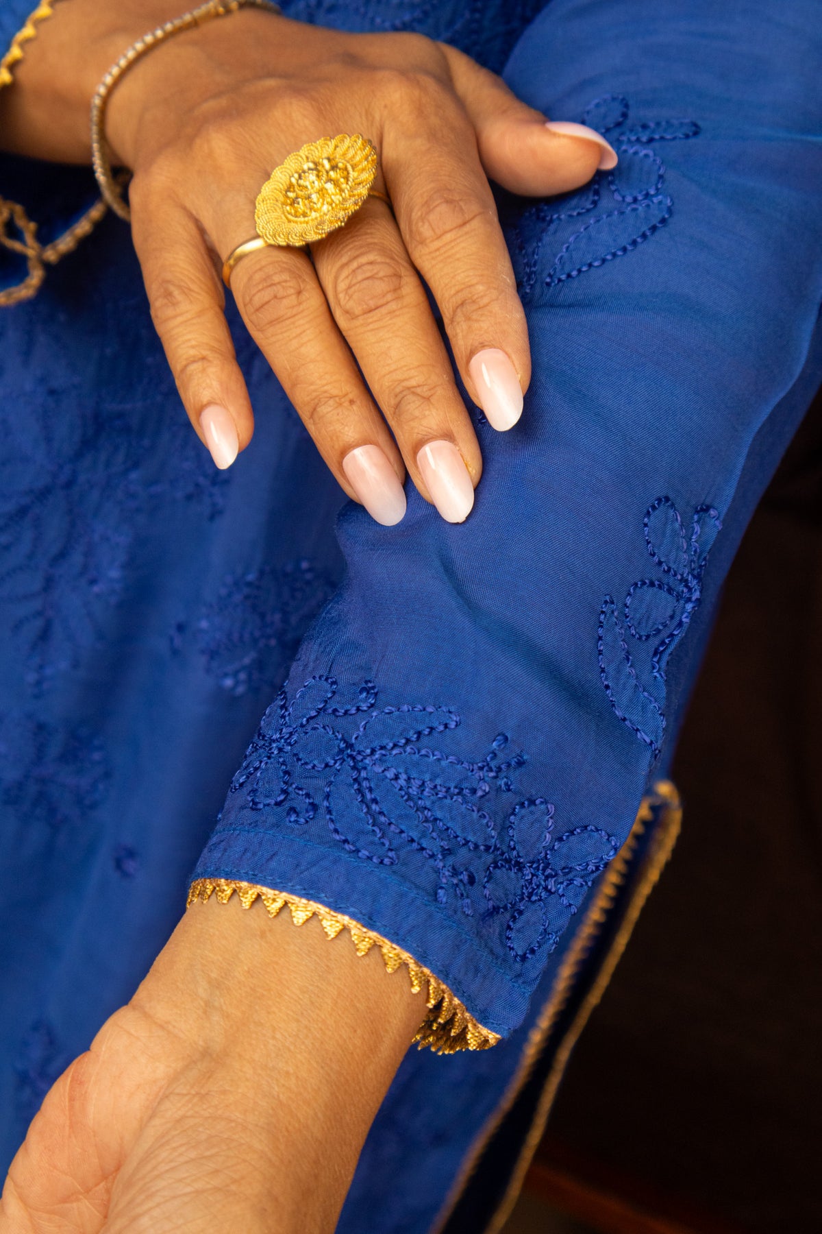 A close-up of a person's hand resting on a royal blue embroidered fabric sleeve, featuring a large textured gold ring and a gold bracelet.