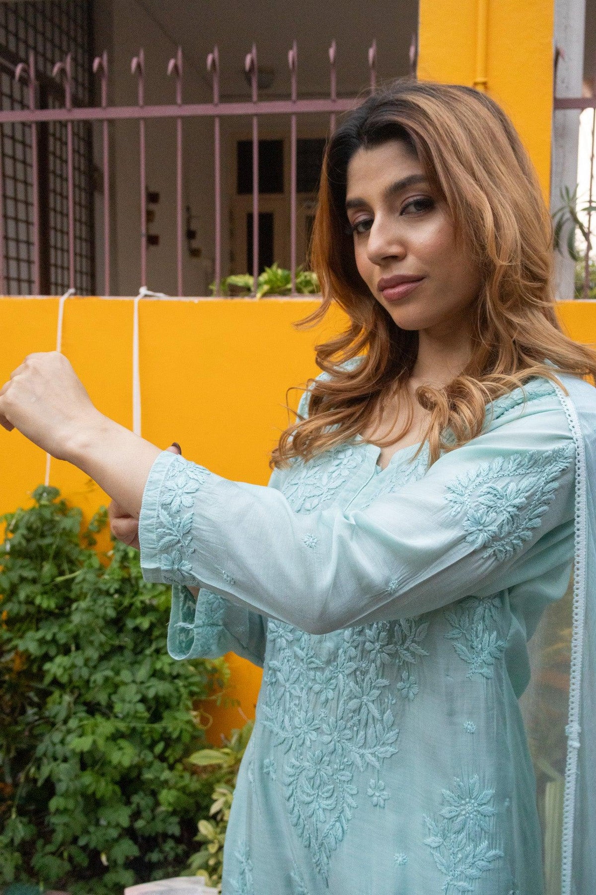 A woman wearing a light blue embroidered traditional outfit, holding a string against a bright yellow wall with green plants in the background. Blue Muslin Chikankari Ghaaspatti Long Kurti with Tone-on-Tone Embroidery