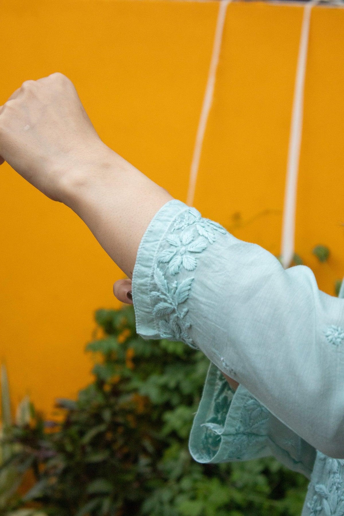 A close-up of a person's forearm wearing a light blue-green embroidered sleeve, set against a bright orange wall and blurred green foliage. Blue Muslin Chikankari Ghaaspatti Long Kurti with Tone-on-Tone Embroidery