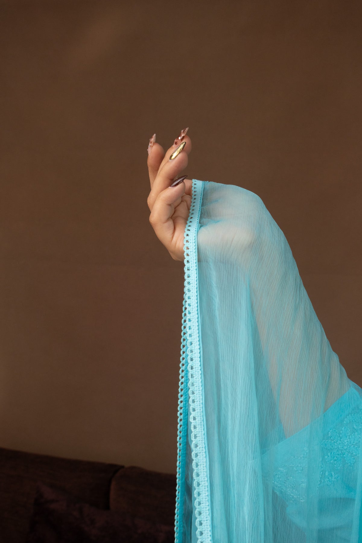 A close-up of a person's hand holding up a sheer light blue fabric drape with a decorative embroidered border, set against a plain brown background.