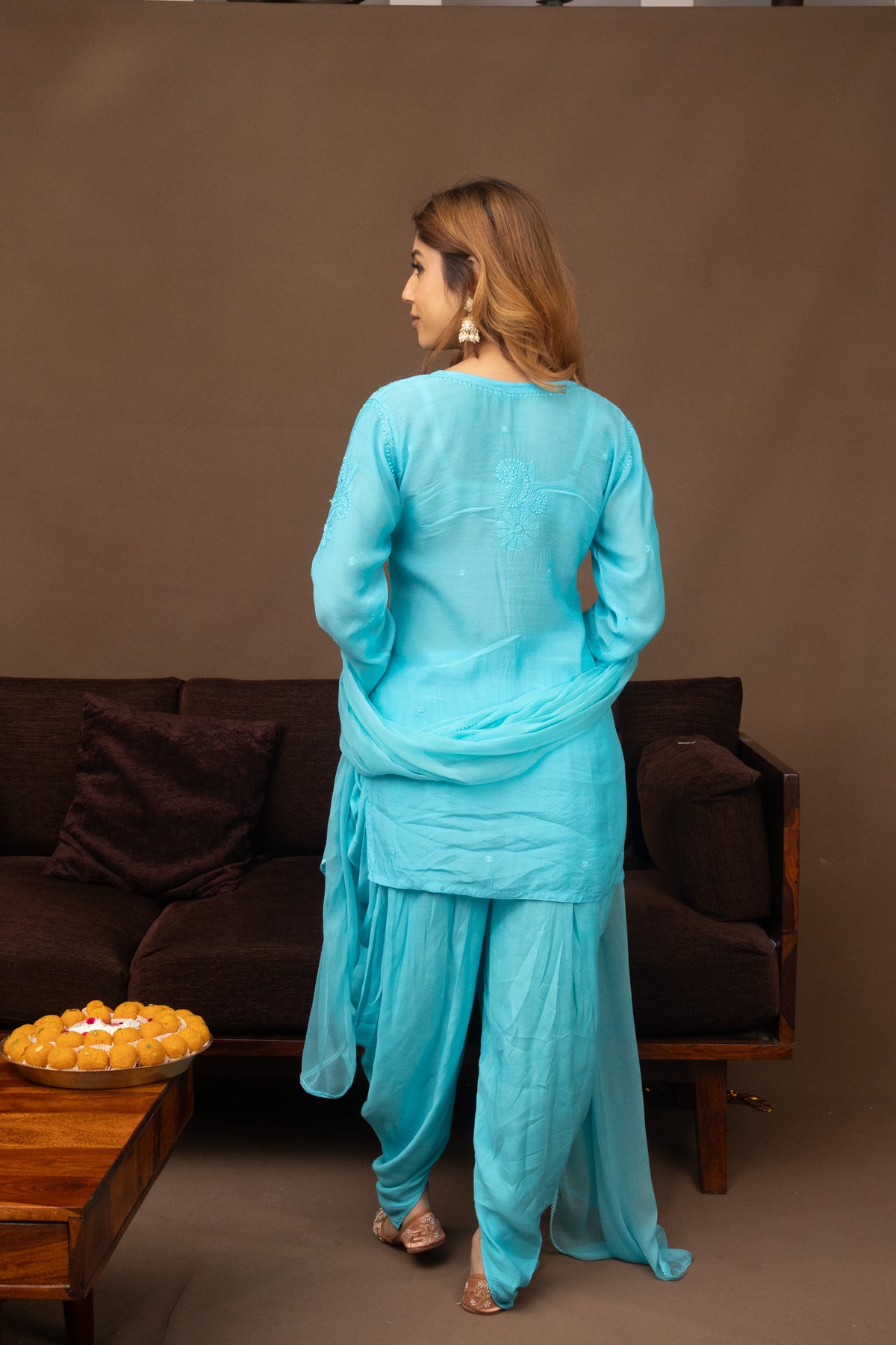 A woman wearing a light blue traditional three-piece salwar kameez suit with matching dupatta, shown from the back in a living room setting with a brown sofa and coffee table.