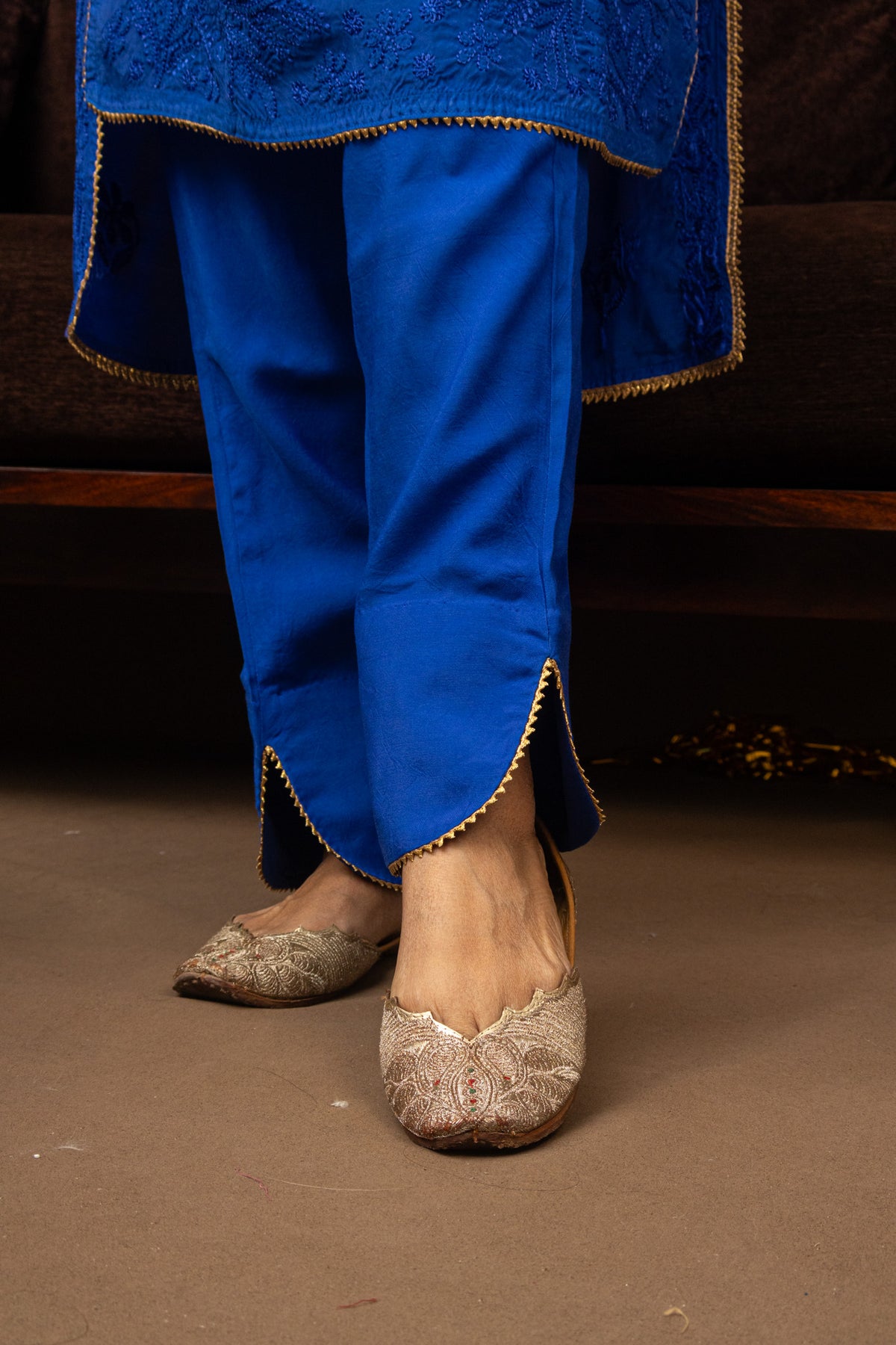 A close-up of a person wearing bright royal blue traditional pants with gold trim and matching blue fabric draped at the hem, paired with beige embroidered flat shoes, standing on a brown floor.
