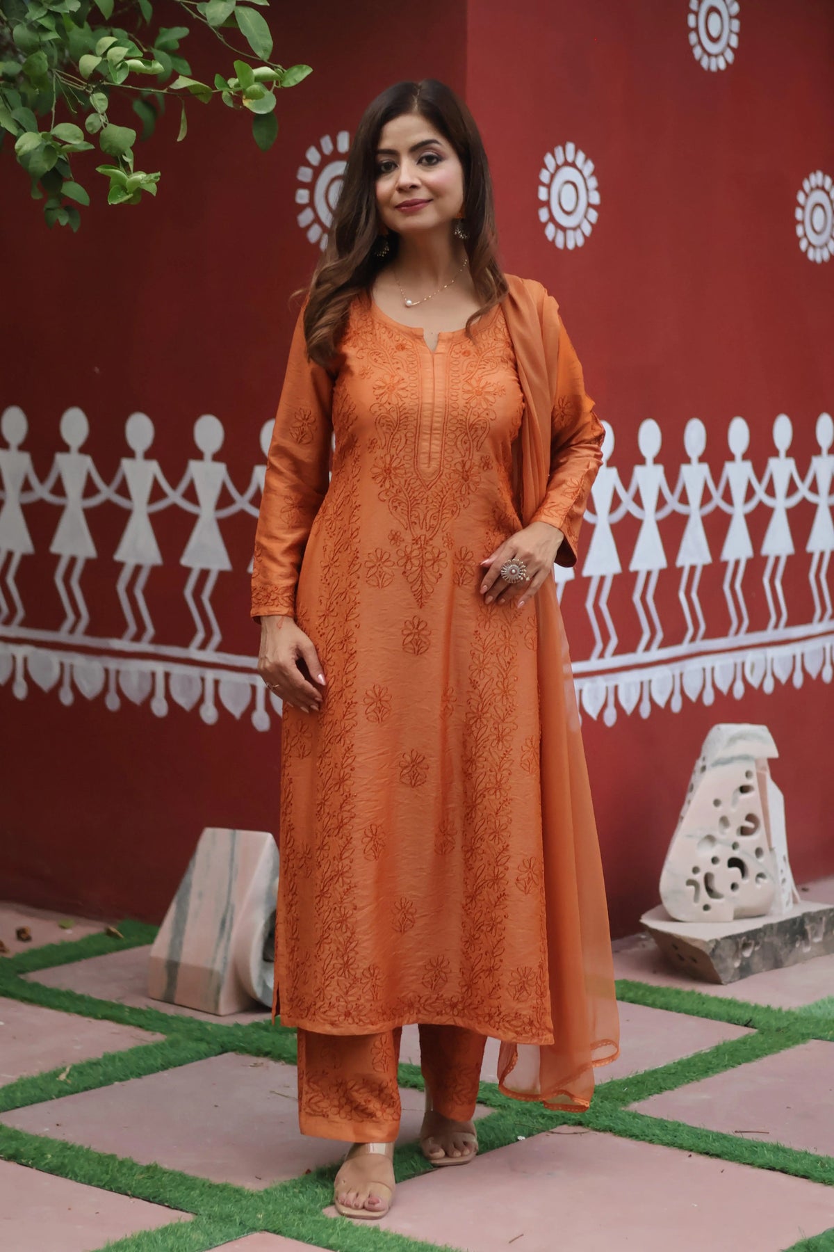 A woman modeling a traditional Indian salwar kameez set in terracotta orange, featuring an embroidered kurta, matching trousers, and a sheer dupatta, set against a red wall with white folk art patterns. Brown Chanderi Chikankari Bakhiya 3 Piece Set with Dupatta Brown Chanderi Chikankari Bakhiya 3 Piece Set with Dupatta 