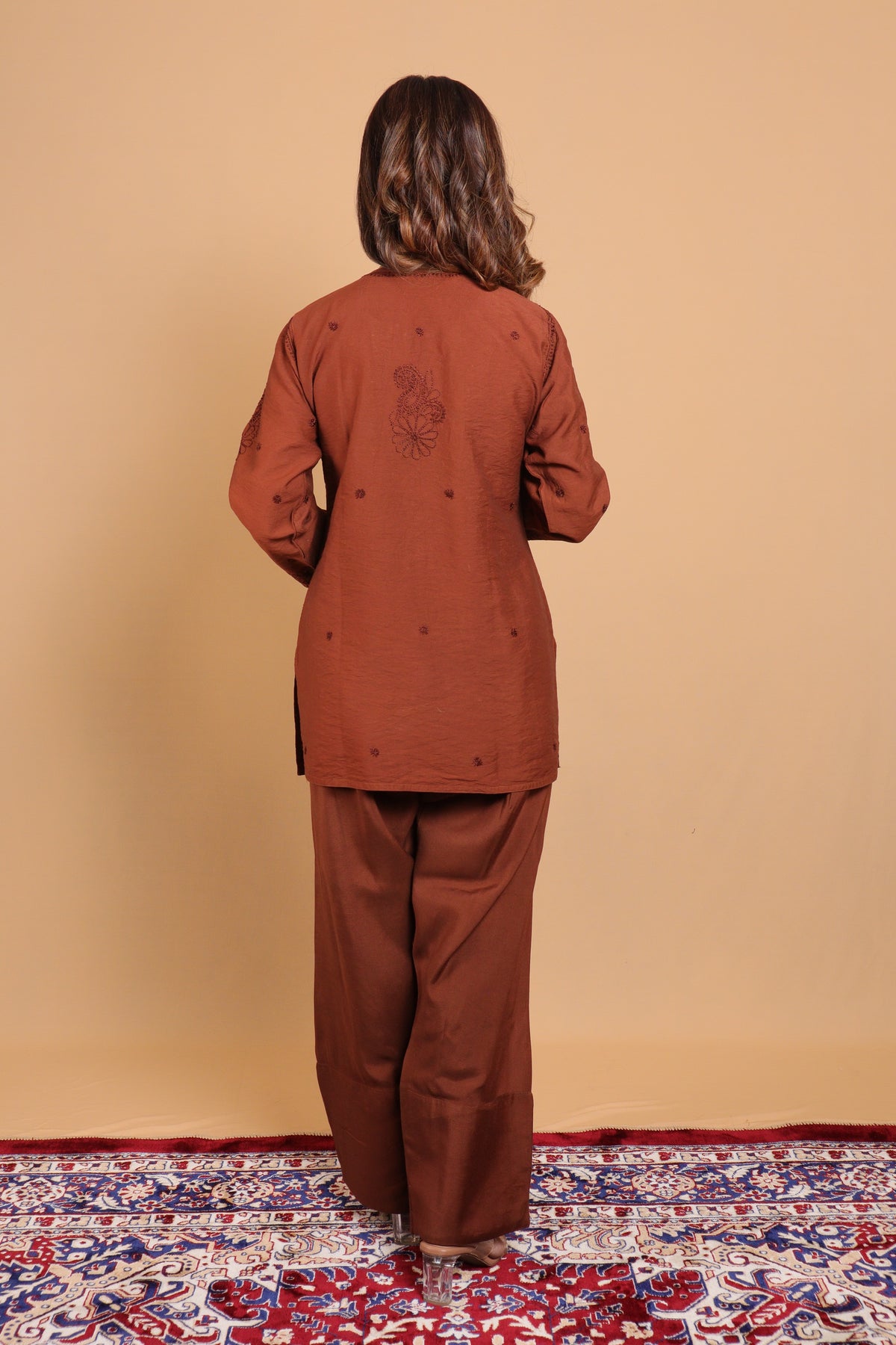 A woman viewed from behind wearing a rust-brown two-piece outfit with a long-sleeve top and wide-leg pants, standing on a patterned red and blue rug against a solid beige wall.