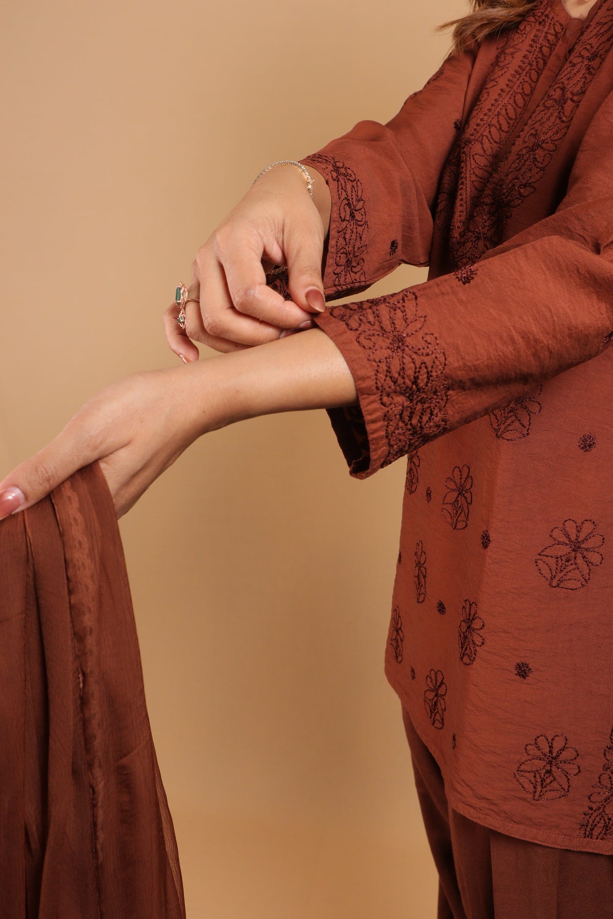 A close-up of a person wearing a rust-brown traditional embroidered kurta with matching pants and a dupatta, shown against a solid beige background.