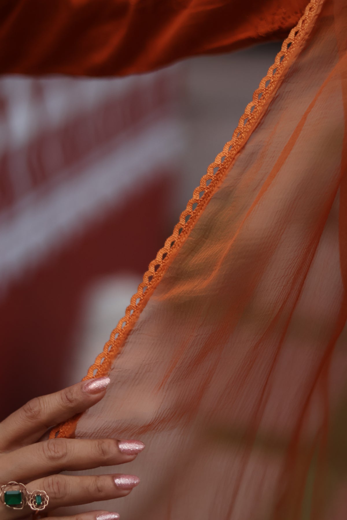 Close-up of a sheer orange fabric with a decorative orange lace trim, held by a hand with pink glitter nail polish and rings.