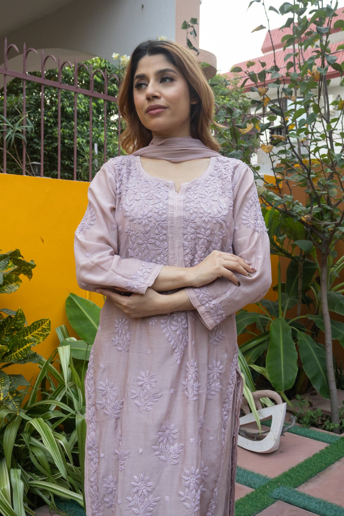 A woman wearing a light pink embroidered kurta with a matching dupatta, standing in a garden setting with a yellow wall and green plants in the background.