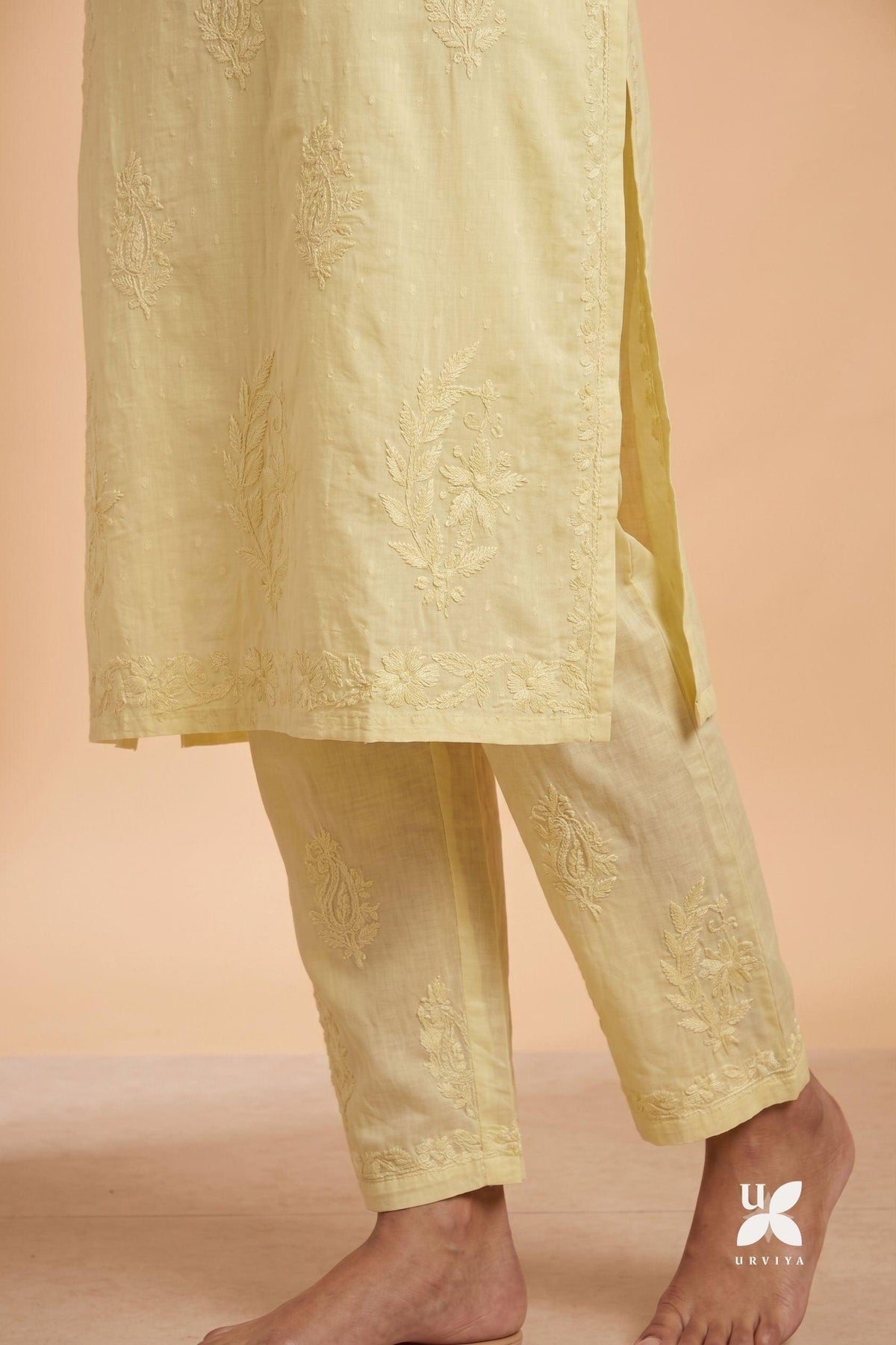 A close-up of a person wearing a light yellow embroidered kurta and matching trousers, shown from the waist down against a warm beige studio background. The fabric has subtle floral embroidery, and bare feet are visible.