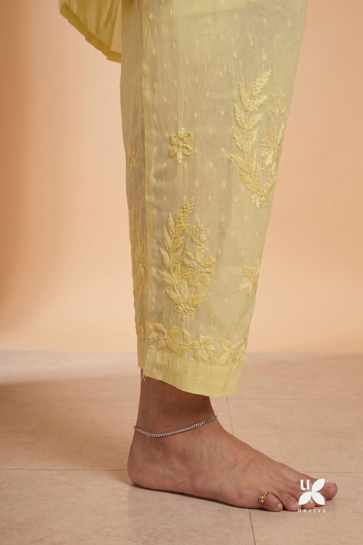 Close-up of a person's bare foot and ankle wearing light yellow embroidered pants with floral and leaf embroidery, set against a warm beige background.
