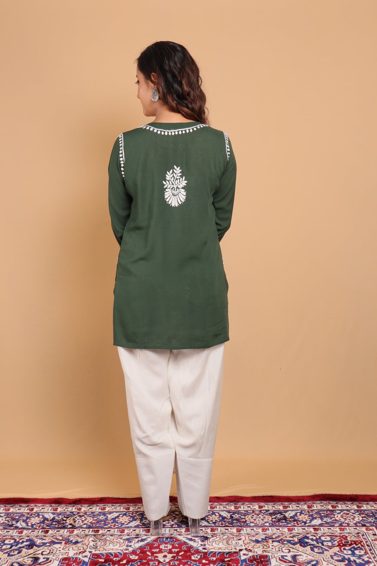 A woman stands with her back to the camera wearing a dark green long-sleeve kurta with white embroidery on the back and white pants, set against a solid beige studio backdrop and a patterned rug.