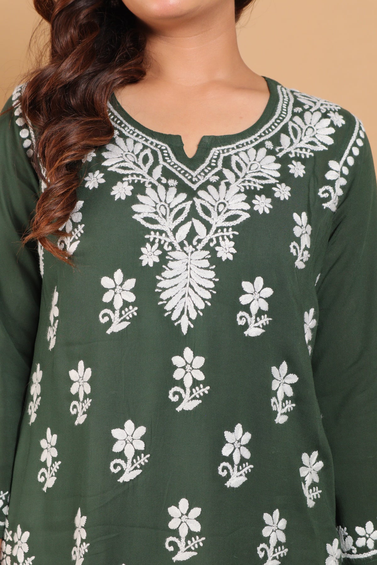 A close-up of a woman wearing a dark green long-sleeve tunic top with white floral and leaf embroidery around the split neckline and scattered across the front, set against a solid beige background.