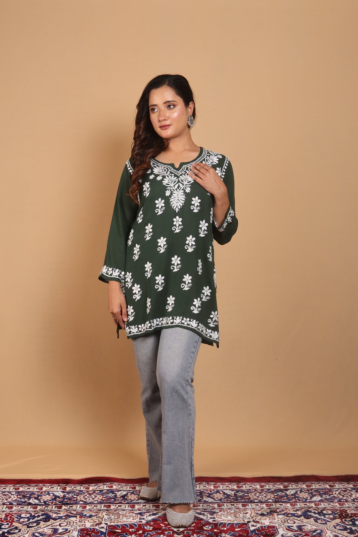 A woman modeling a dark green kurta with white floral embroidery, paired with light gray jeans, standing against a solid beige backdrop.