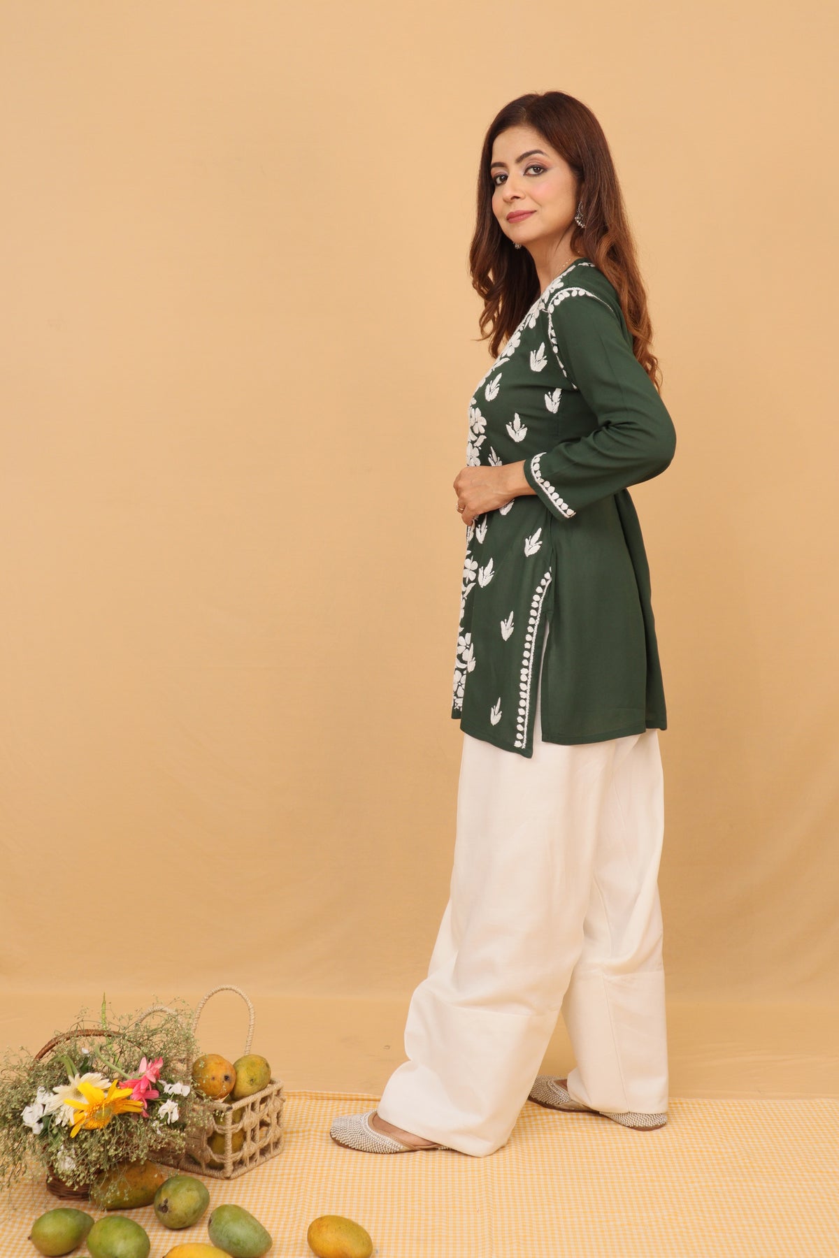 A woman modeling a dark green long-sleeve kurta with white printed motifs and white wide-leg pants, standing against a solid beige backdrop with a small decorative basket and fruit props at the bottom left.