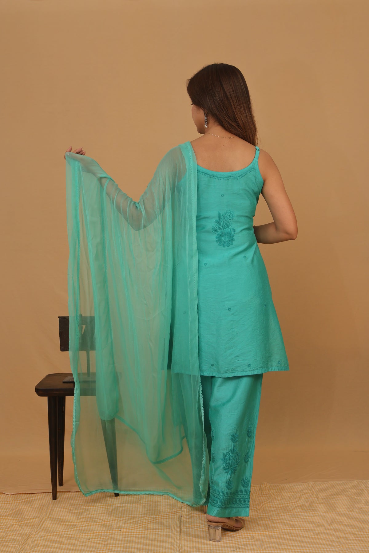 A woman stands with her back to the camera wearing a turquoise green ethnic three-piece suit with a matching sheer dupatta, set against a solid beige studio background.