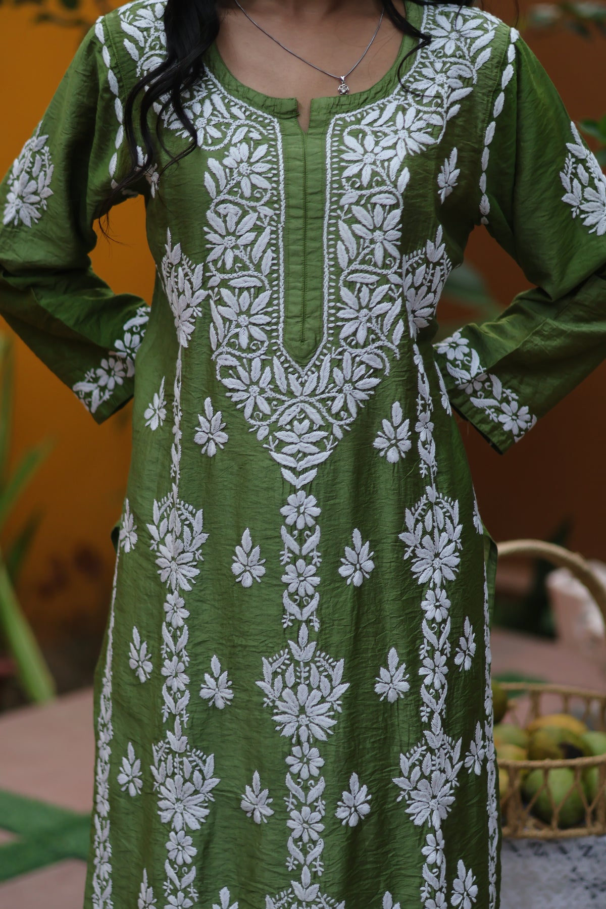 A woman wearing a green long kurta with white floral embroidery and printed patterns, standing against a warm orange-brown background.