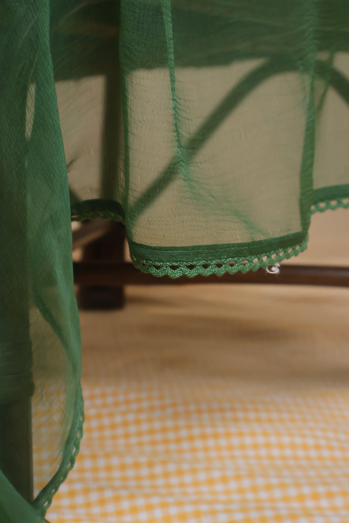 A close-up of a green sheer mesh fabric with a scalloped lace trim, draped over a table in an indoor setting with a yellow-and-white checkered surface below. Green Chiffon Dupatta -17 with Crochet Lace