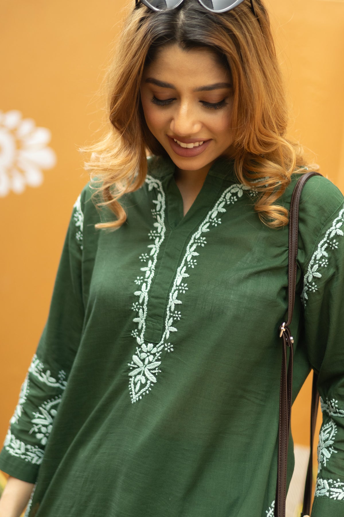 A woman wearing a dark green cotton kurta with white floral embroidery on the neckline and sleeves, set against a solid orange background.