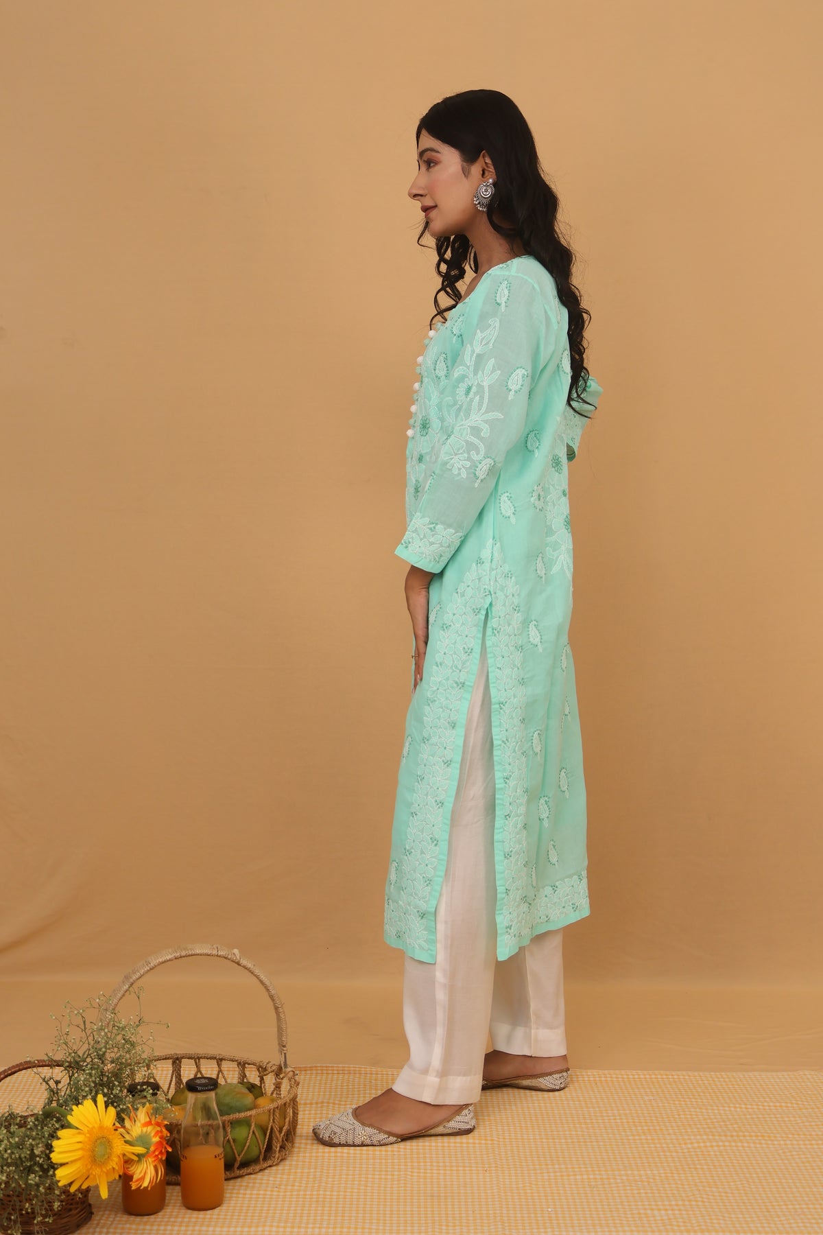 A woman stands in profile wearing a mint green embroidered kurta with white pants, posed against a solid beige studio backdrop with a small decorative basket and flowers at her side.
